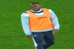 Ein Füßballspieler in einem orangen T-Shirt und blauen Hosen überquert entschlossen ein grünes Feld.