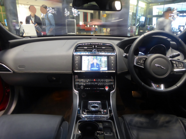 Innenansicht eines Jaguar F-Pace R-Dynamic S AWD mit Lenkrad, Armaturenbrett, Bildschirm, Knöpfen, Sitzen, Spiegel und Glasfenstern. Personen, Lichter und andere Gegenstände sind durch die Fenster zu sehen.