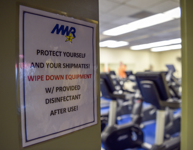 Ein Schild an einer Turnhalle mit der Aufschrift "Schütze Dich und Deine Schiffskameraden" mit Laufbändern und Deckenleuchten im Hintergrund, das Bild leicht unscharf, um Bewegung anzudeuten.