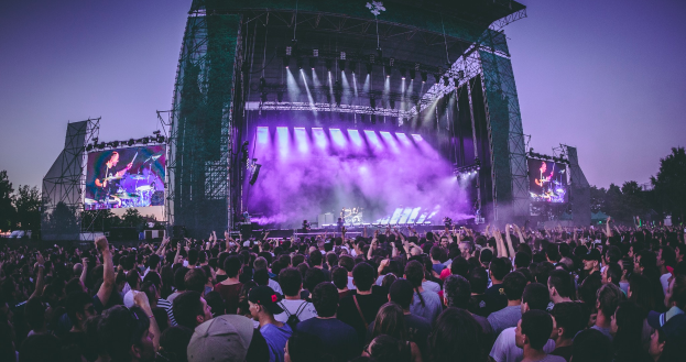 Eine große Menschenmenge auf einem Musikfestival, mit einer Bühne in der Mitte, auf der Musiker Instrumente spielen, beleuchtet von Lichtern und umgeben von Bildschirmen, unter einem klaren blauen Himmel mit einem Gebäude und Bäumen im Hintergrund.