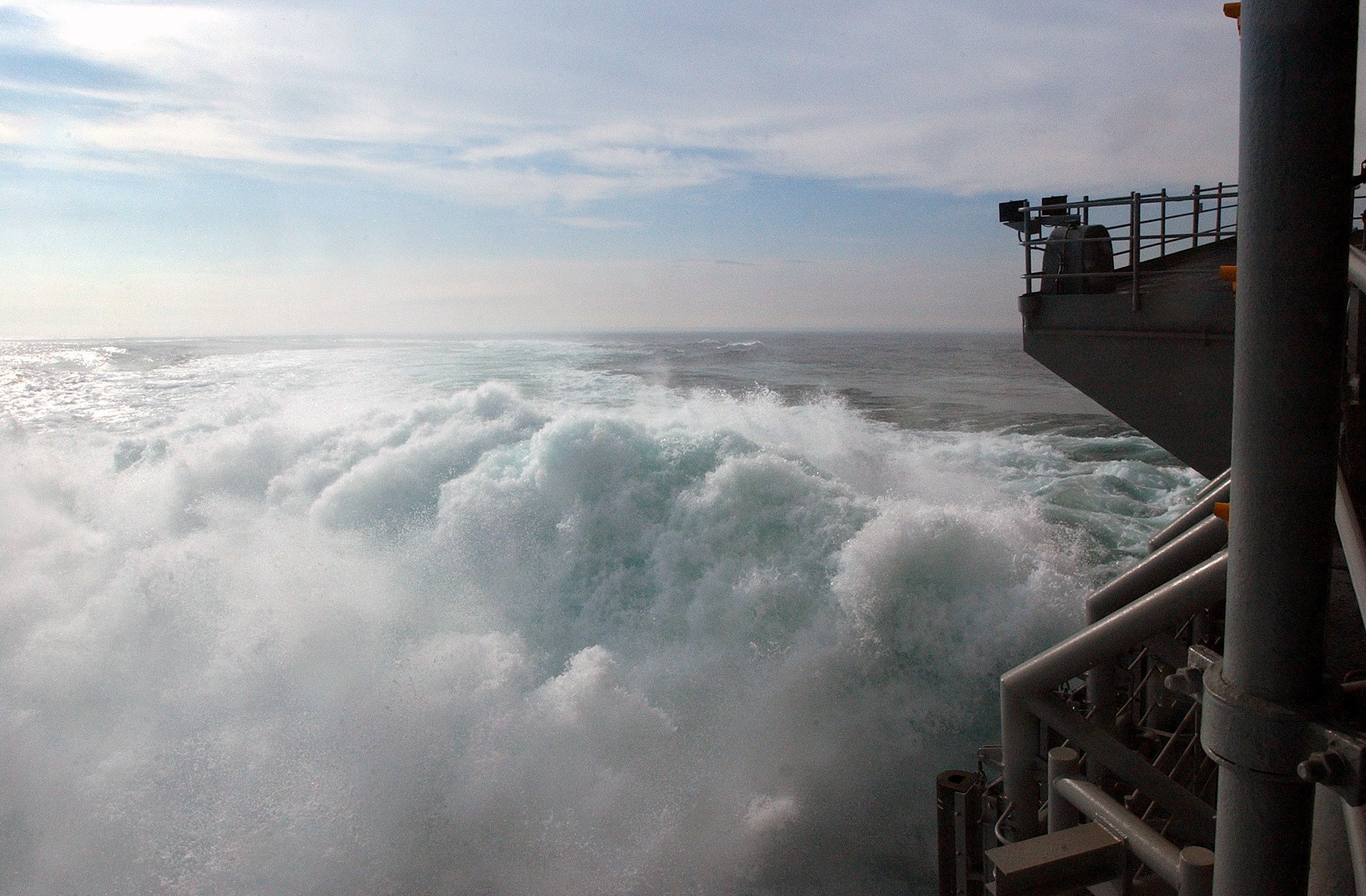 Große Welle, die auf das Deck eines Schiffs kracht, mit sichtbaren Relings und Pfählen
