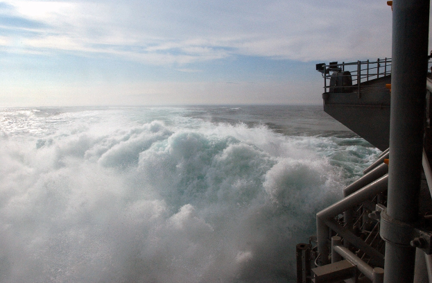 Große Welle, die auf das Deck eines Schiffs kracht, mit sichtbaren Relings und Pfählen