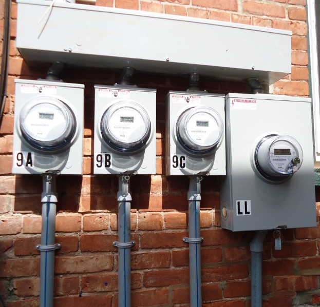 Drei Stromzähler an der Seite eines Backsteingebäudes montiert, verbunden mit Rohren, mit einem Fenster auf der rechten Seite.