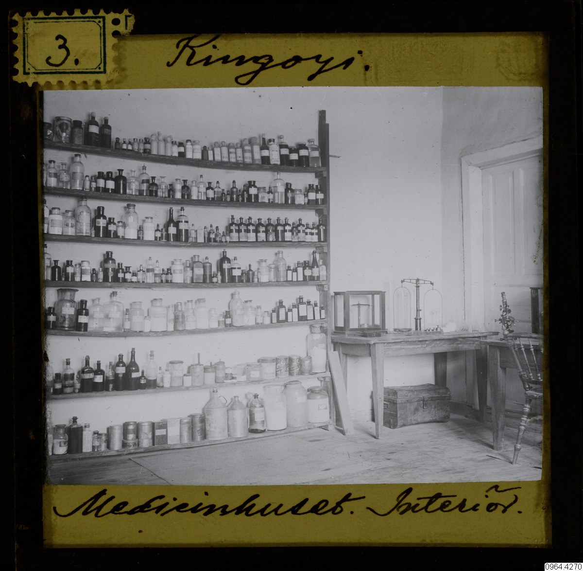 Schwarz-weißes Foto eines alten Apothekeninneren mit Regalen voller Flaschen, einem Tisch mit Gegenständen, einem Stuhl, einer Tür und einer Wand, mit Text unten.