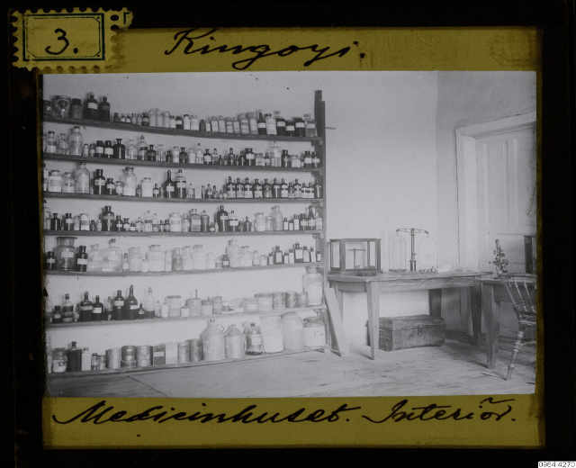 Schwarz-weißes Foto eines alten Apothekeninneren mit Regalen voller Flaschen, einem Tisch mit Gegenständen, einem Stuhl, einer Tür und einer Wand, mit Text unten.