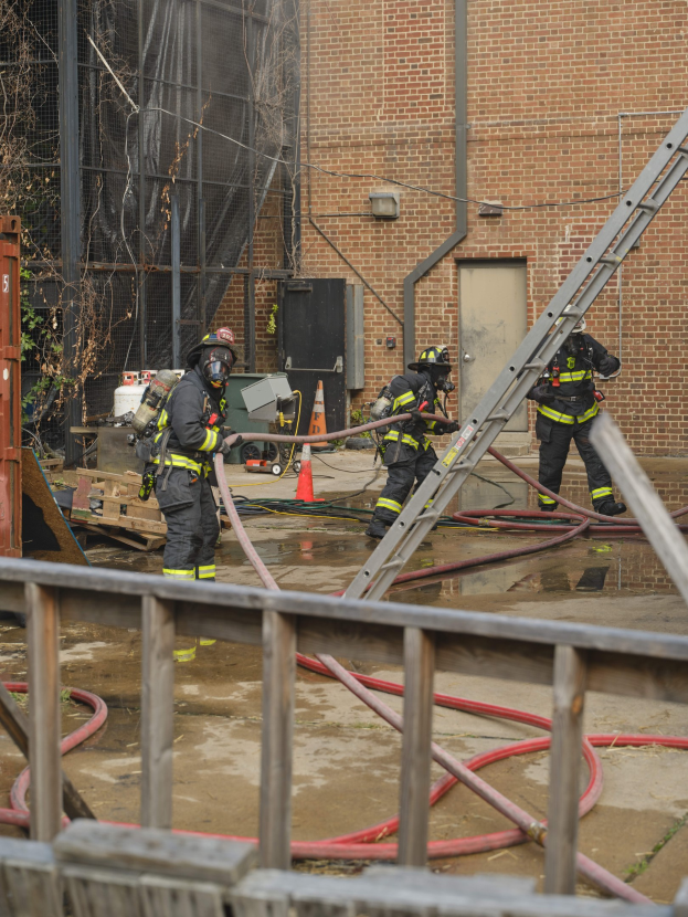 Feuerwehrleute in Helmen arbeiten daran, ein Gebäude Feuer zu löschen, während sie Schläuche halten, mit einem Metallzaun, verstreuten Rohren, einem Behälter, einem Verkehrskegel und Hintergrundelementen wie einem Baum und Himmel.