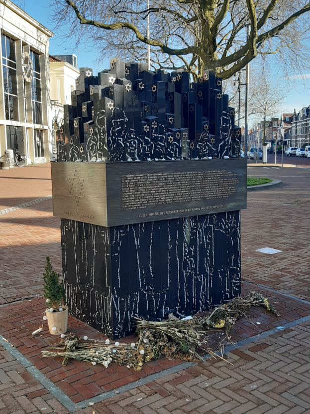 Denkmal für Holocaust-Opfer in einem Stadtplatz, mit einem Baum davor, einer Topfpflanze, verstreuten trockenen Blättern und Gebäuden, Fahrzeugen und Bäumen im Hintergrund.