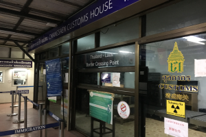 Der Eingang eines Zollamts in Bangkok, Thailand, mit Glaswänden und -türen, Informationsschildern mit Text und Bildern, Pfählen mit Bändern, einem Schild, einem Dach mit Deckenleuchten und einer Wand mit verschiedenen Gegenständen.