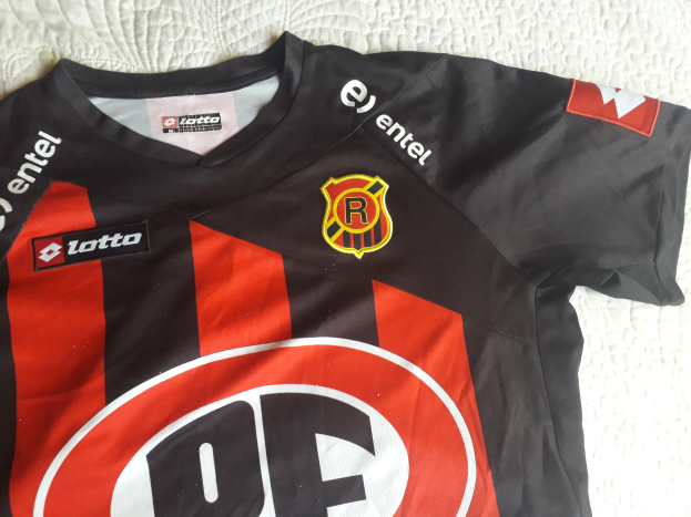 Ein schwarzes und rotes Fußballtrikot mit einem roten und schwarzen Streifen, einem Logo und Text, das auf einer weißen Oberfläche liegt.