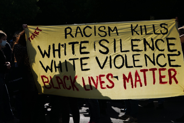 Eine Gruppe maskierter Menschen steht nachts auf einer Straße und hält ein Schild hoch, auf dem steht: "Rassismus tötet Weiße Stille, weiße Gewalt, Schwarze Leben zählen."