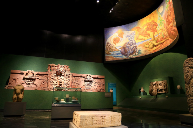 Ein Museumsraum mit verschiedenen Kunstwerken, darunter Skulpturen, Tischobjekte, eine Texttafel, eine Wandmalerei und Deckenlampen im Mexiko-Stadt-Museum für Anthropologie.
