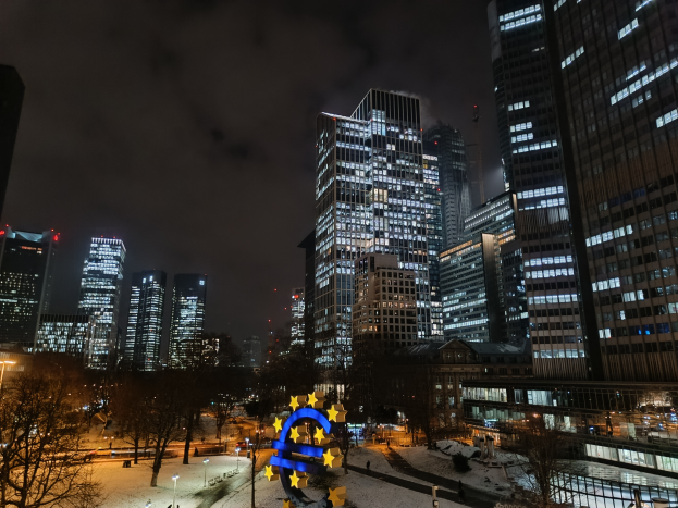 Das Hauptquartier der Europäischen Zentralbank in Frankfurt, Deutschland bei Nacht, mit beleuchteten Gebäuden, Bäumen, Straßenlaternen, Straßenlaternen und Menschen auf der Straße, unter einem bewölkten Himmel, mit einem Wasserzeichen auf dem Bild.