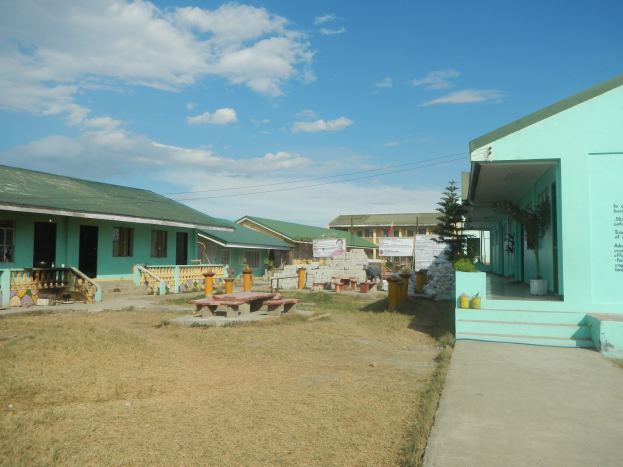 Ein Schulgebäude mit grüner Dachfläche und blauem Himmelhintergrund, das Fenster, Türen, Treppen, Geländer, eine Treppe, Bänke, Topfpflanzen, Texttafeln, Bäume und Grasfläche mit verstreuten Wolken zeigt.