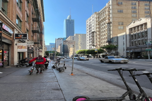 Eine Gruppe von Menschen, die auf Rollern eine städtische Straße mit hohen Gebäuden entlangfahren, mit einem geparkten Pick-up, Straßenlaternen, sichtbarem Himmel, Bäumen und Schildern.