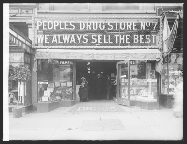 Ein Schwarz-Weiß-Foto eines Drugstores mit einem Schild, auf dem "People's Drug Store No. 7 - Wir verkaufen immer das Beste" steht, mit ein paar Menschen drinnen und einem Topfpalme links daneben.