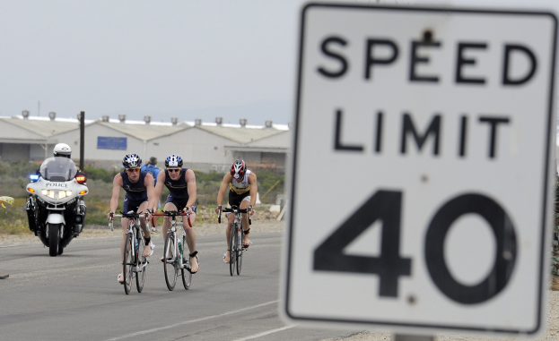 Gruppe von Radfahrern in Helmen auf einer Straße mit einem Geschwindigkeitsbegrenzungsschild, Gebäude und Bäume im Hintergrund unter einem klaren blauen Himmel.