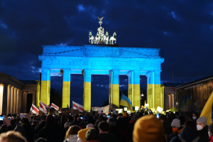 Menschenmenge mit Fahnen und Schildern vor dem Brandenburger Tor, mit einem Banner auf der rechten Seite mit protestrelevanten Text.