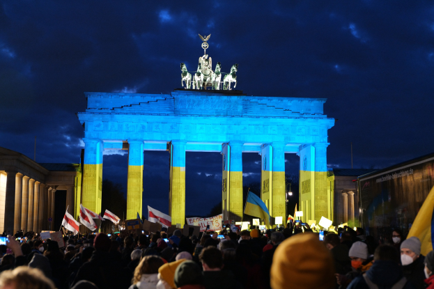 Menschenmenge mit Fahnen und Schildern vor dem Brandenburger Tor, mit einem Banner auf der rechten Seite mit protestrelevanten Text.