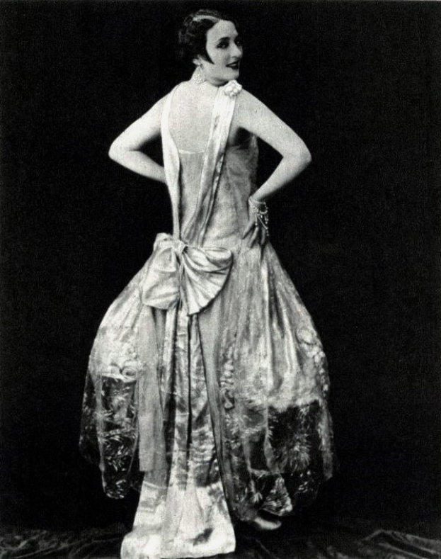 Eine Frau in einem Kleid im Stil der 1920er Jahre mit hohem Halsausschnitt, langen Ärmeln und einem weiten Rock steht selbstbewusst, ihre Haare sind elegant frisiert und das Make-up natürlich.