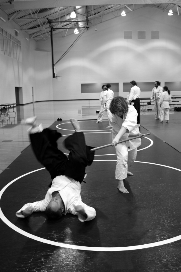 Zwei Personen in einer Turnhalle üben Karate mit Stöcken, umgeben von anderen, mit beleuchteten Wänden im Hintergrund.