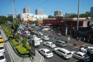 Eine belebte Stadtstraße mit verschiedenen Fahrzeugen, Fußgängern, Gebäuden und städtischer Infrastruktur unter einem klaren blauen Himmel.
