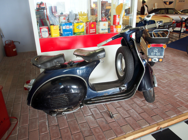 Ein schwarzer und weißer Roller steht vor einer Tankstelle, daneben ein rotes Objekt und ein Feuerlöscher sowie mehrere Fahrzeuge, eine Person und Schilder im Hintergrund.