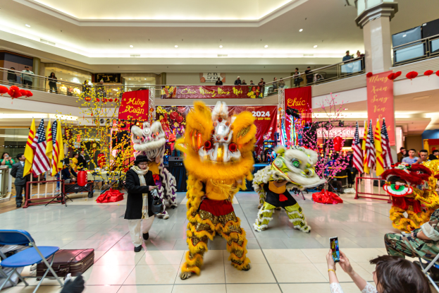 Ein lebendiges chinesisches Neujahrsfest in einem Einkaufszentrum mit Menschen in Kostümen, die tanzen, und anderen, die auf Stühlen sitzen und mobile Telefone halten, umgeben von Flaggen, Bannern, Dekorationen und architektonischen Elementen mit einem Wasserzeichen unten.
