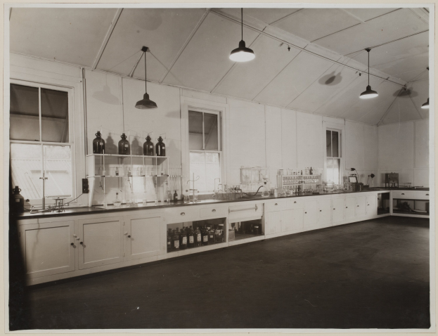 Schwarzes und weißes Foto eines Labors mit einer langen Arbeitsfläche, Schäubern, Regalen mit Flaschen, Fenstern, Deckenleuchten und verschiedenen Gegenständen auf der Arbeitsfläche.