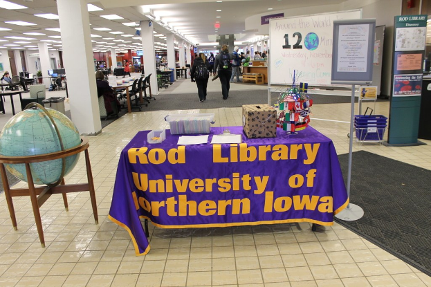 Expo-Stand mit Flaggen verschiedener Länder, einer Box, Papieren und einem Banner auf einem Tisch, einer Kugel links daneben, einer Mitteilungstafel mit Menschen in der Nähe rechts daneben und zusätzlichen Tischen und Stühlen im Hintergrund.