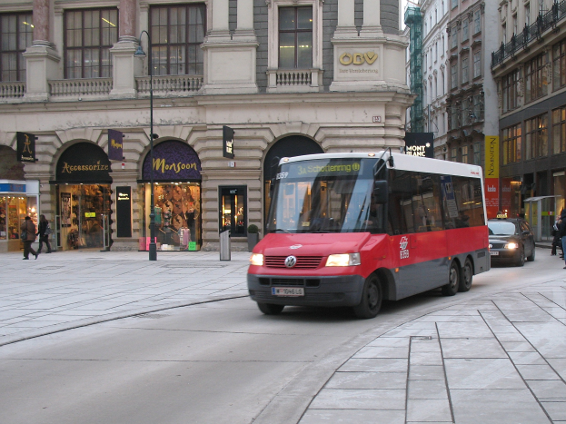 Roter Shuttlebus fährt auf einer Stadtstraße mit hohen Gebäuden, Passanten auf dem Gehweg, Laternen und Schaufenstern im Hintergrund unter einem sichtbaren Himmel.