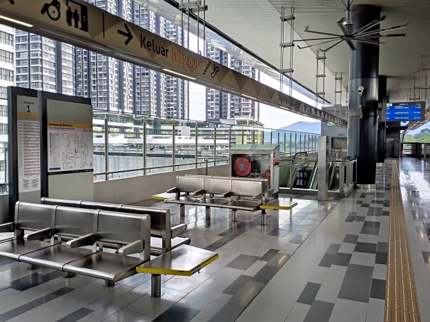 Leerer Flughafen Wartebereich mit mehreren Bänken, Informationsanzeigen, Säulen, Deckenventilator und Deckenleuchten, vor einem Hintergrund aus Gebäuden, Bäumen, Wasser und einem klaren blauen Himmel.