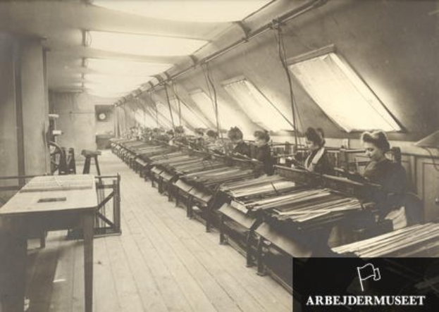 Schwarz-weiß-Foto von Frauen, die Textilmaschinen in einer Fabrik bedienen, mit Tischen links und einer Uhr an der Wand im Hintergrund.