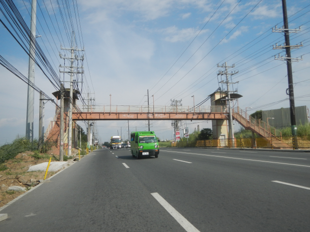 Ein grüner Lkw fährt auf einer Straße neben einer Brücke, mit Strommasten und -leitungen an der Straße und Bäumen, Gebäuden und einem klaren blauen Himmel im Hintergrund.
