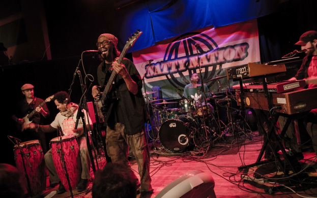 Eine Live-Band performt auf der Bühne mit Mikrofonen und Ständern, während das Publikum darunter unter einer dunklen und mysteriösen Atmosphäre mit einem Banner im Hintergrund zusieht.