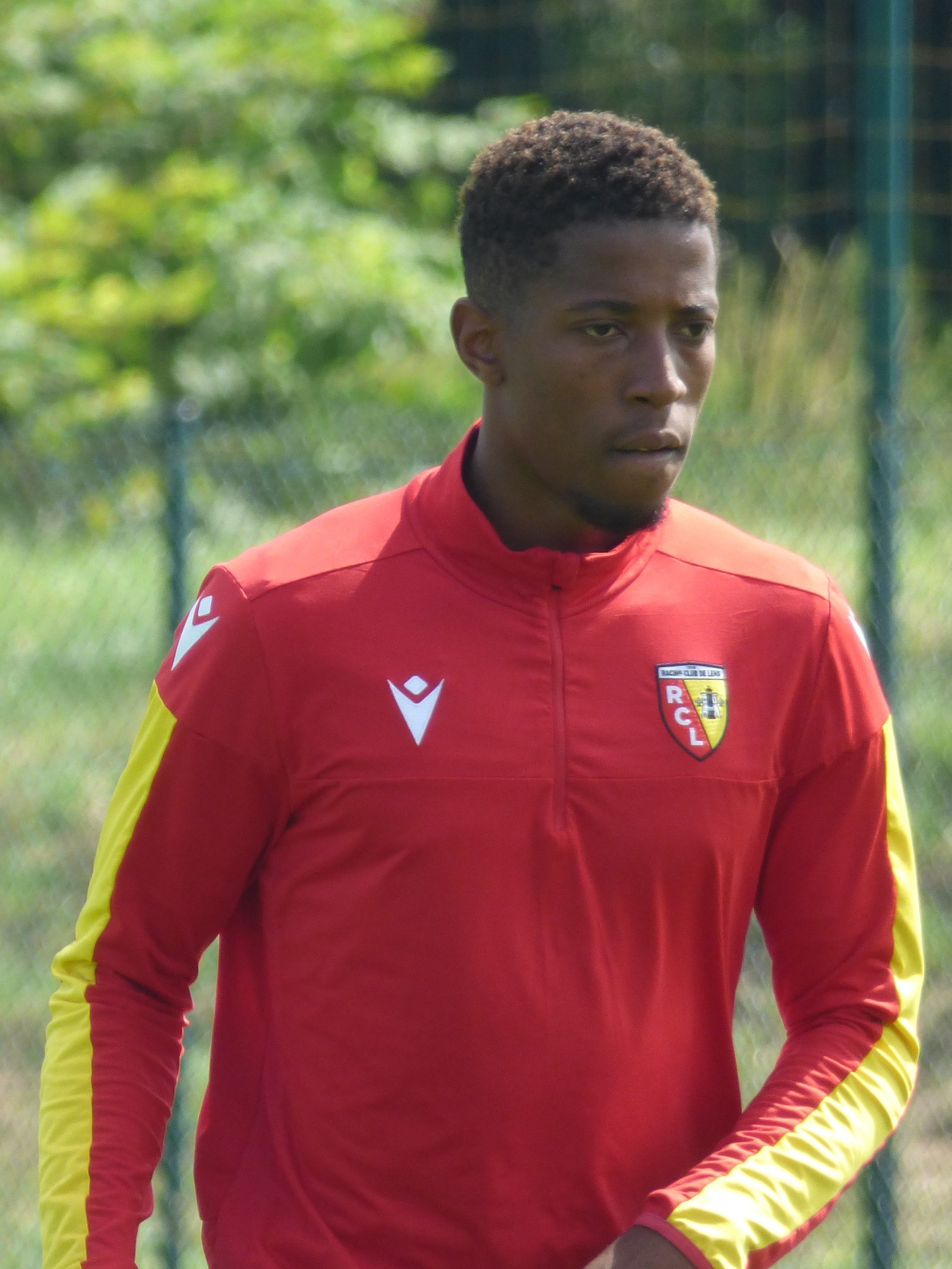 Ein junger Mann in einem roten und gelben Trikot steht auf einem Fußballfeld, das von einem Zaun und Bäumen umgeben ist und sieht entschlossen aus.