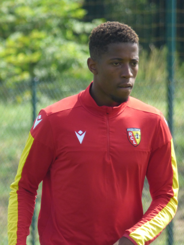 Ein junger Mann in einem roten und gelben Trikot steht auf einem Fußballfeld, das von einem Zaun und Bäumen umgeben ist und sieht entschlossen aus.