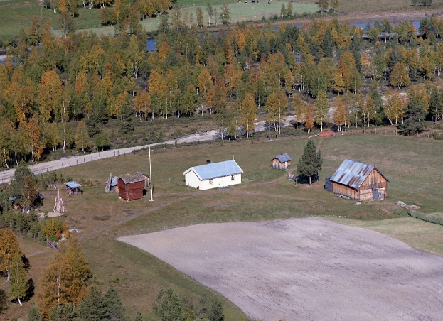 Luftaufnahme eines Ranchgeländes mit verstreuten Gebäuden in einem Feld umgeben von Bäumen, Häusern, Strommasten, einer Straße, Gras und Wasser.