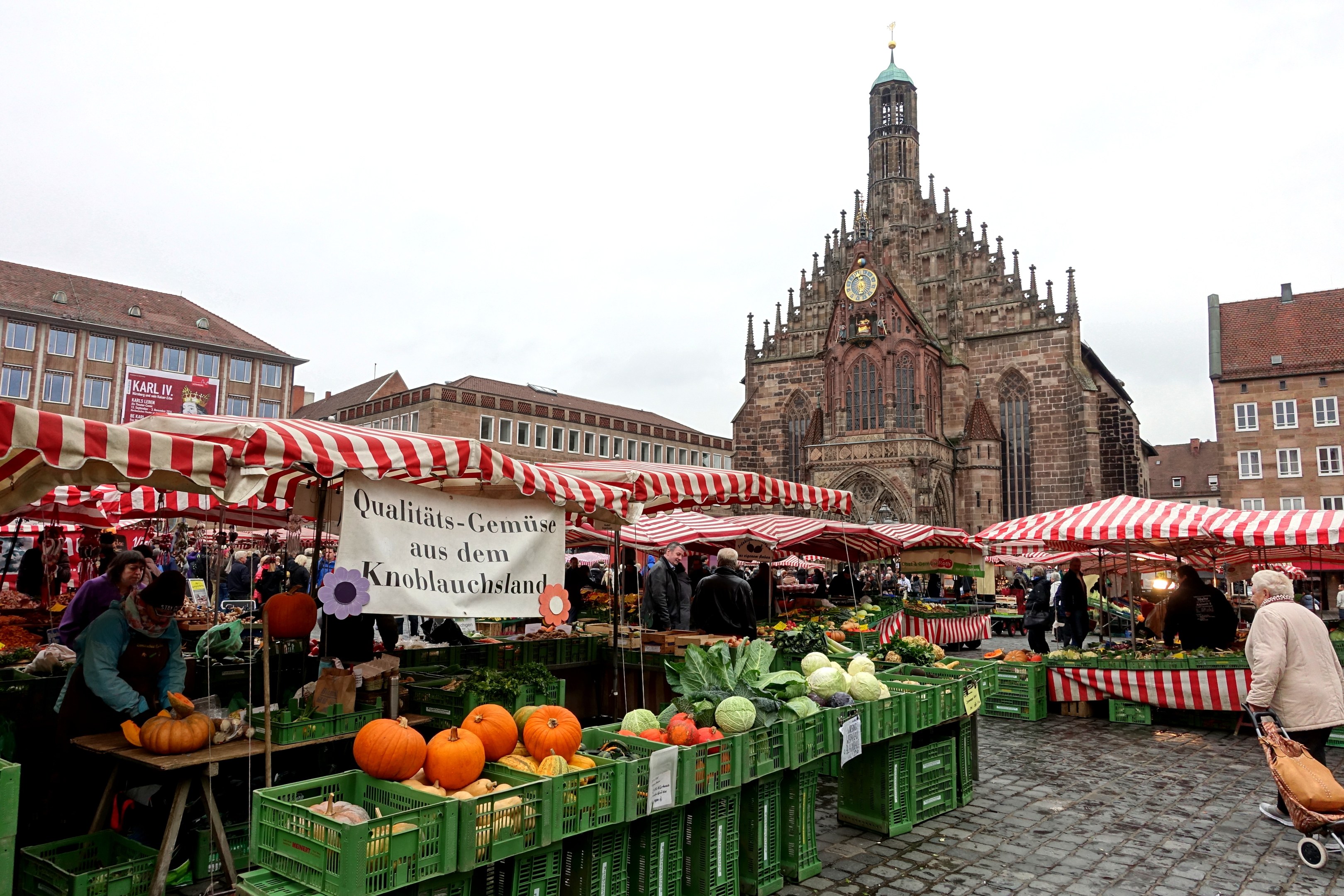 Ein belebter Markt in Nürnberg, Deutschland, mit farbenfrohen Obst- und Gemüsesorten, Menschen mit Taschen und Zelten, vor einem Hintergrund aus Gebäuden mit Fenstern und einem Uhrenturm unter einem sichtbaren Himmel.