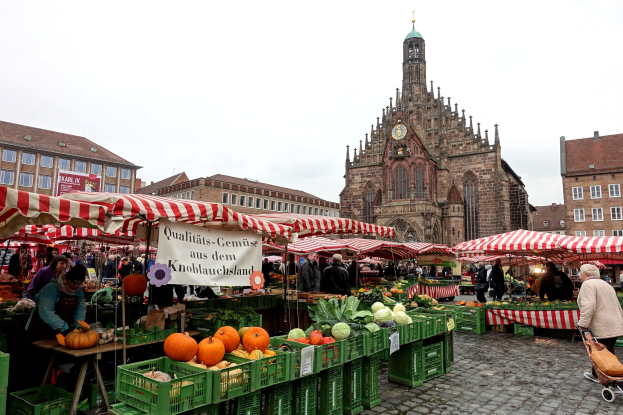Ein belebter Markt in Nürnberg, Deutschland, mit farbenfrohen Obst- und Gemüsesorten, Menschen mit Taschen und Zelten, vor einem Hintergrund aus Gebäuden mit Fenstern und einem Uhrenturm unter einem sichtbaren Himmel.