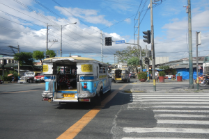 Ein blauer und weißer Bus fährt auf einer Straße neben einer Ampel, umgeben von Strommasten mit Kabeln, Laternen, Bäumen, Gebäuden und einem bewölkten Himmel.