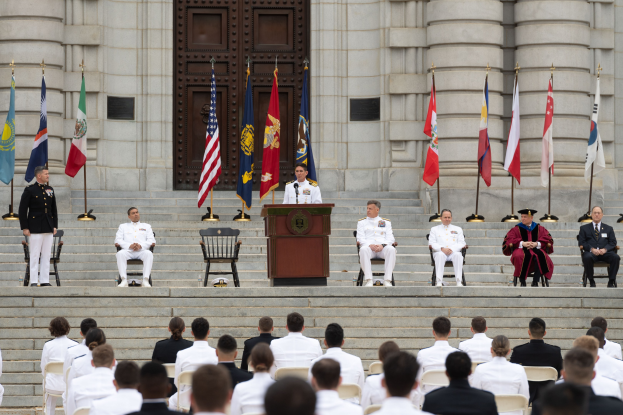 Gruppe von Menschen in weißen Uniformen auf einer Treppe sitzend mit einem Podium, das von Fahnen flankiert ist, vor einem großen Gebäude mit einer Tür.