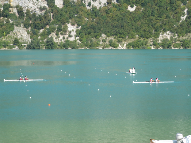 Eine Gruppe von Menschen, die auf einem Gewässer mit einem bewaldeten Hügel im Hintergrund und einem Boot am Bildrand rudern, was auf ein Rennen hindeutet.