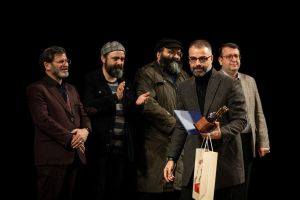 Eine Gruppe von Männern steht auf einer Bühne mit einem, der eine Tasche und eine Trophäe hölt, und feiert die Eröffnung der Berliner Filmfestspiele vor einem dunklen Hintergrund.