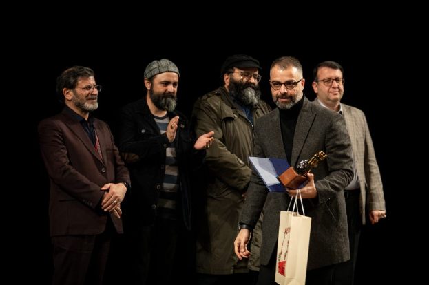 Eine Gruppe von Männern steht auf einer Bühne mit einem, der eine Tasche und eine Trophäe hölt, und feiert die Eröffnung der Berliner Filmfestspiele vor einem dunklen Hintergrund.