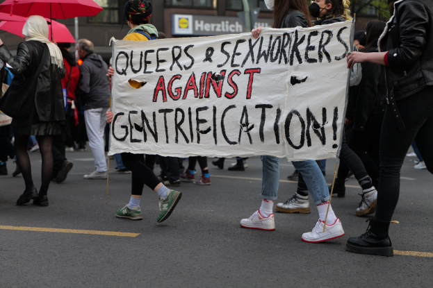 Gruppe von Menschen marschiert mit einem Banner, auf dem "Queers und Sexarbeiter gegen Gentrifizierung" steht, einige tragen Masken und einer hält einen Schirm, mit Bäumen und einem Gebäude im Hintergrund.