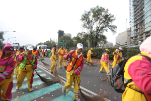 Eine Gruppe von Menschen in gelben Uniformen, Mützen und mit Taschen, die die Straße mit Besen kehren, während Fahrzeuge vorbeifahren, mit Bäumen, Gebäuden, Laternenmasten und einem klaren blauen Himmel im Hintergrund.