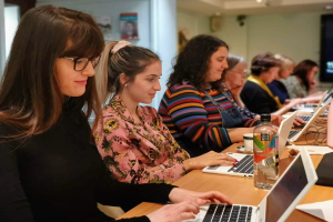 Eine Gruppe von Frauen, die um einen Tisch mit Laptops und Bürobedarf arbeiten, was auf einen Online-Kurs hindeutet, in einem Raum mit einem Foto, einer Säule und Deckenleuchten.