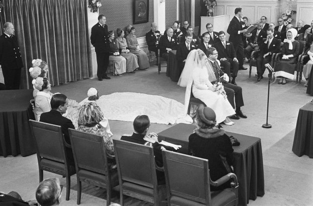 Schwarz-weiß-Foto einer Hochzeitszeremonie mit sitzenden und stehenden Gästen, einem Mann mit Mikrofon, einer Frau mit Blumen, einem Podium mit Mikrofon, Blumenschmuck, einem Vorhang, einem gerahmten Foto an der Wand und einer Tür im Hintergrund.