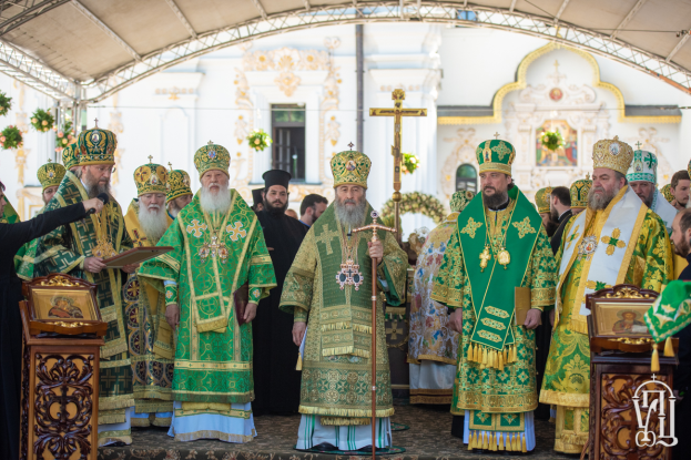 Eine Gruppe von Priestern vor einer Kirche, wobei einer ein Buch und ein Mikrofon auf der linken Seite hält und ein Kreuz, Blumen und ein Gebäude im Hintergrund zu sehen sind.