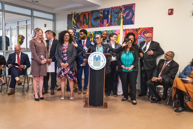 Gruppe von Menschen, die vor einem Rednerpult mit Mikrofon und einer Tafel mit dem Text 'Sancho City Councilman Joins City Council' stehen, mit sitzenden und stehenden Zuschauern, Flaggen, einem Feuerlöscher und Deckenleuchten im Hintergrund.